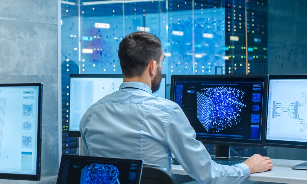 A man sitting at a desk and looking at three computer screens that display charts and graphs from a network monitoring tool.