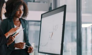 Two young businesswomen looking at an interactive whiteboard to analyze investment data, representing private equity and venture capital.